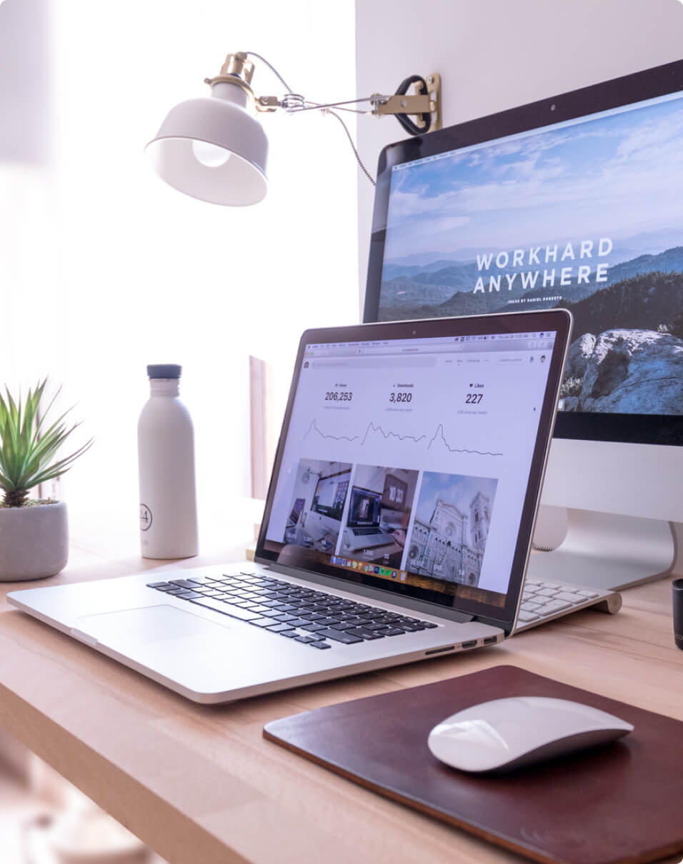 Apple laptop and MacBook open on a wooden desk with a lamp, plant, mouse and water bottle.