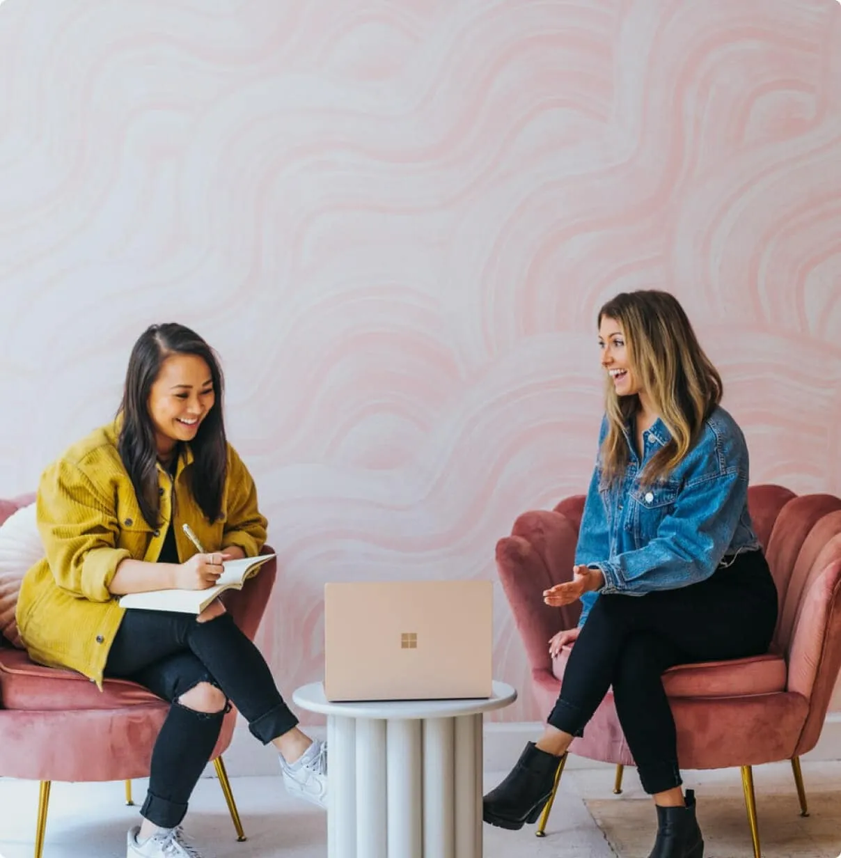 Two women sitting in pink chairs having a friendly discussion with a laptop on the table between them. One is writing in a notebook while the other gestures animatedly.