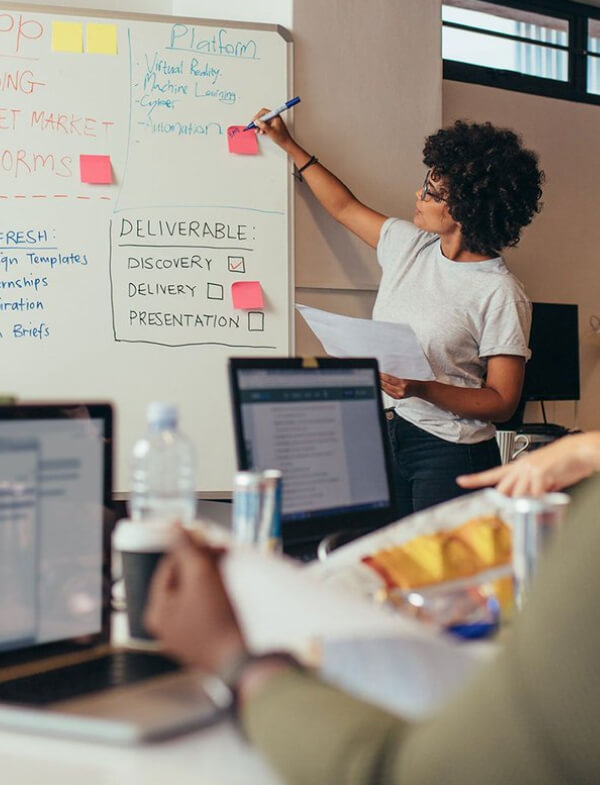 A woman standing in front of a whiteboard filled with notes listening to, and writing down, suggestions from co-workers.