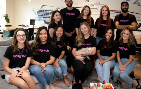 A group of twelve people, some seated and some standing, pose indoors wearing matching black "linegraph" t-shirts; one person is holding a small dog.