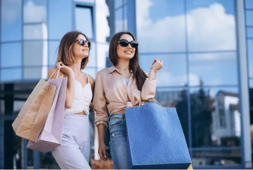 Two stylish young women wearing sunglasses are happily walking with colorful shopping bags in hand, standing outside a modern glass-fronted shopping mall on a sunny day.