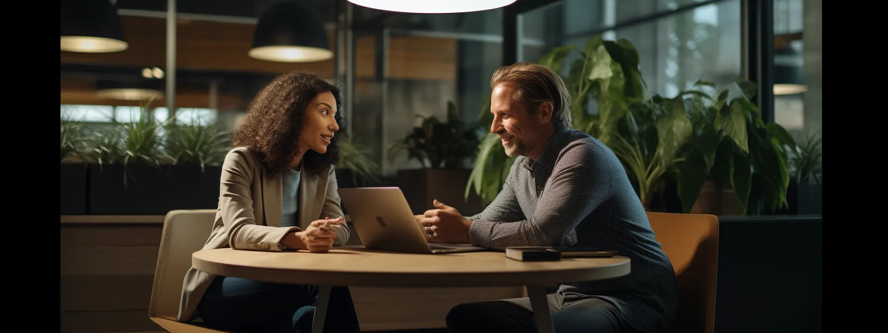 Two people sit at a round table in a modern office, having a discussion. One uses a laptop while the other listens attentively. Indoor plants and large windows are in the background.