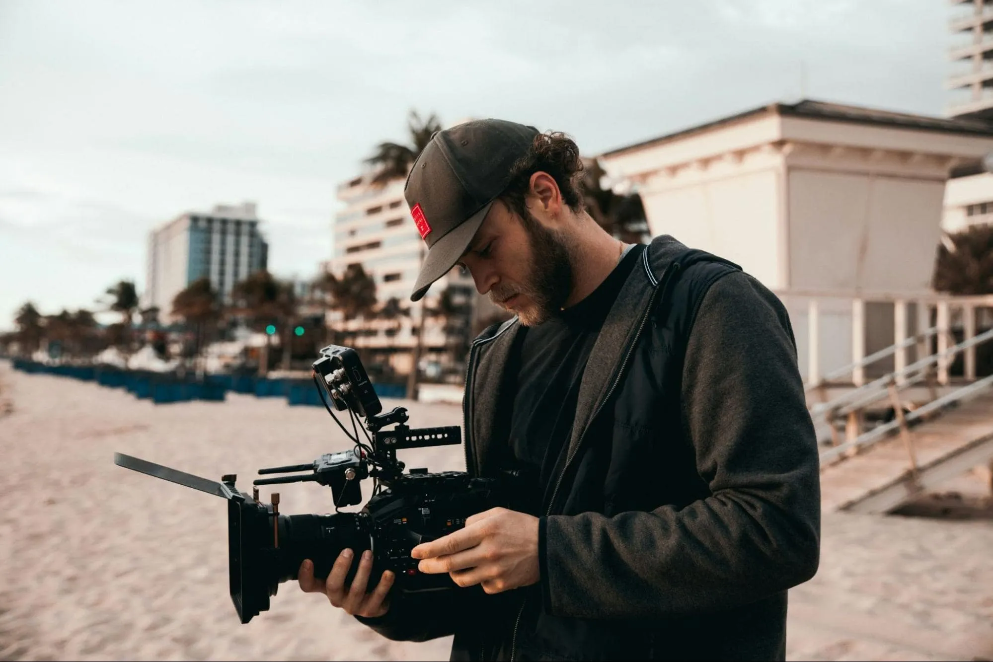 A man on the beach checking a professional video camera, with modern buildings and palm trees in the background.