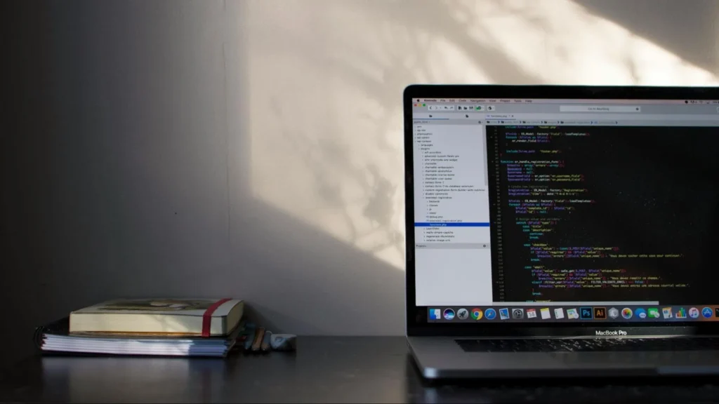 MacBook Pro on a desk displaying PHP code in a dark-themed editor. Next to the laptop is a small stack of books and notebooks, with soft sunlight casting shadows on the wall behind.