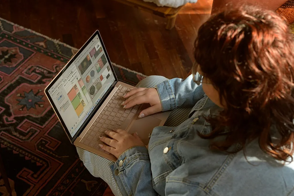 Woman in glasses working on a presentation or design project on a laptop while sitting on the floor with a patterned rug