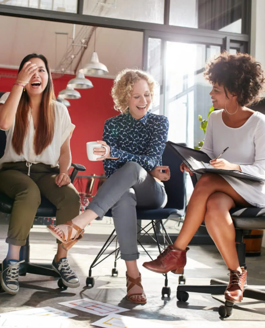 Three diverse women laughing and collaborating in a modern office meeting