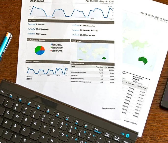 Printed Google Analytics reports with charts and graphs are placed on a wooden desk beside a pen, a partial view of a computer keyboard, and documents referencing Enterprise SEO Software.