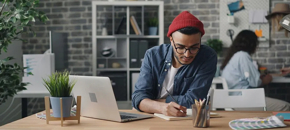 Man wearing glasses and a red beanie writes in a notebook at a desk with a laptop, plants, and office supplies—researching Enterprise SEO Software; another person works in the background.