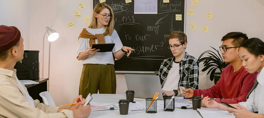 A woman stands and presents to four seated colleagues in a meeting room, discussing strategies with notes, a chalkboard, and Enterprise SEO Software tools in the background.