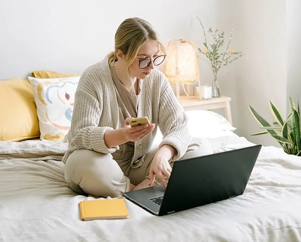A person sits cross-legged on a bed using a laptop and holding a smartphone, with Enterprise SEO Software open on the screen, a notebook nearby, and soft natural lighting in the room.